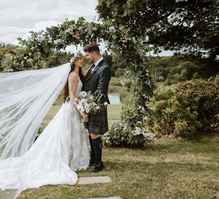 Bride wearing celestial 3d applique sleeveless wedding dress and cathedral length veil blowing in the wind with groom in classic wedding kilt in front of circular floral and foliage arch at The Barn at Harburn wedding venue 