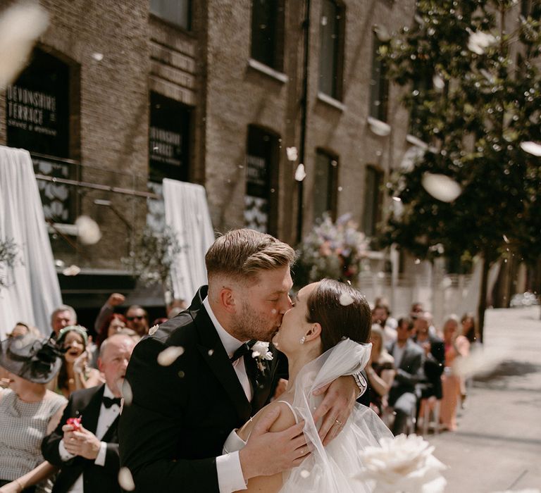 Bride in satin wedding dress with veil kisses the groom in black tux for the first time as a married couple with confetti exploding around them 