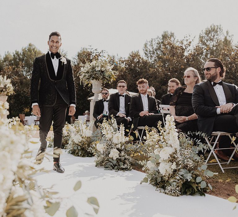 Groom in classic black tuxedo walking down the aisle with carnation, eucalyptus and dried flower aisle flower decorations 