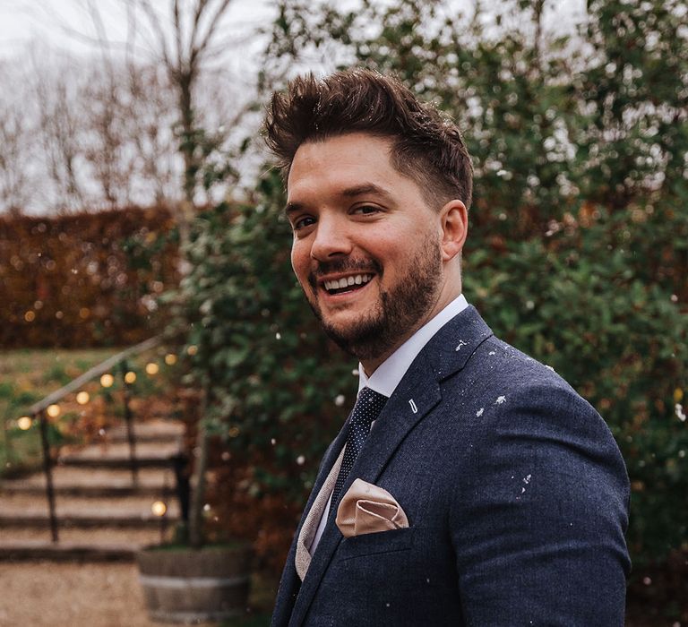 Smiling groom in navy suit with spotted tie and cream waistcoat with watch groom accessory on his wrist walking in the snow on the wedding day