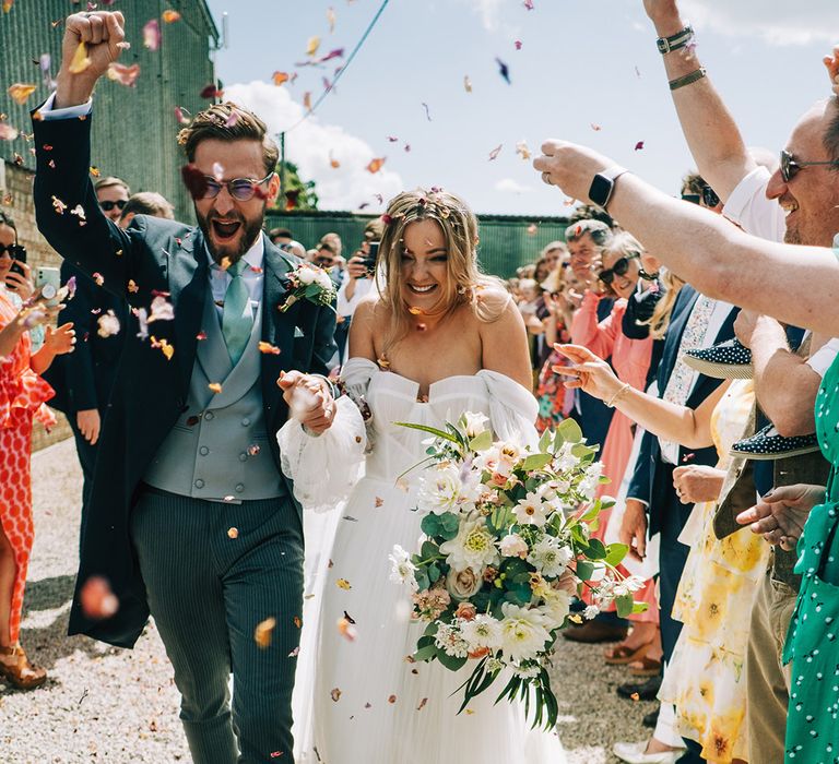 Bride in off the shoulder wedding dress walking with the groom in a morning suit walking to hand-dried rose petal confetti 