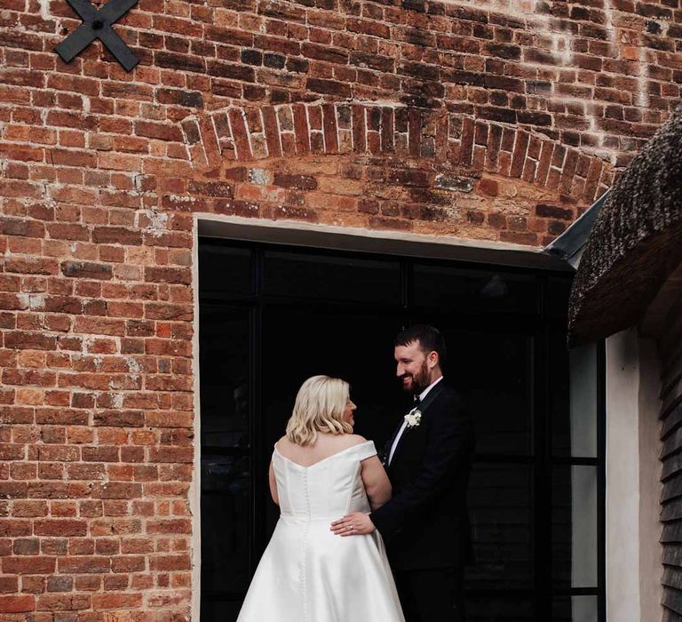 Bride in satin off the shoulder wedding dress standing with groom in classic black tuxedo at Brickhouse Vineyard Devon wedding venue