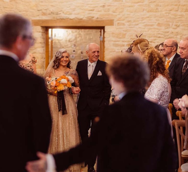 Bride in sleeveless deep v-neck sparkly gold wedding dress with sheer and gold polka dot wedding veil holding orange garden rose, white poppy, light pink carnation and foliage bridal bouquet wrapped in sparkly gold ribbon with father of the bride walking down the aisle at Kingscote Barn Tetbury wedding venue