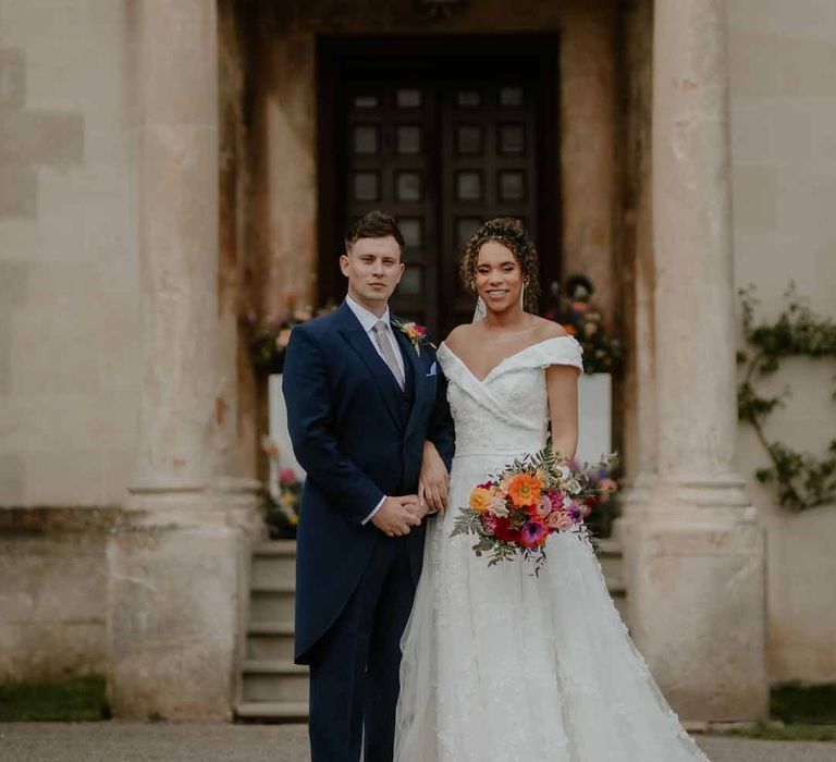 Bride in Suzanne Neville off shoulder 3D floral applique wedding dress standing with groom in navy groom morning suit with grey ties and blush rose boutonniere outside of Elmore Court Gloucestershire wedding