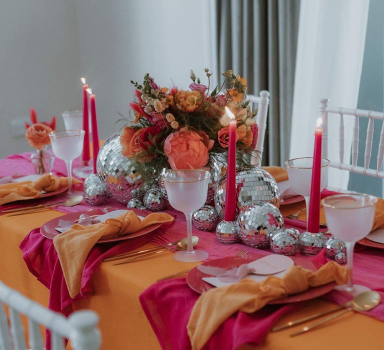 Pink and orange wedding tablescape with orange tablecloth, bright pink wedding table runners, orange wedding napkins, pink tapered candles, pink and orange poppies, garden roses, carnations, dried flowers and foliage wedding table centrepieces and disco ball wedding decor 