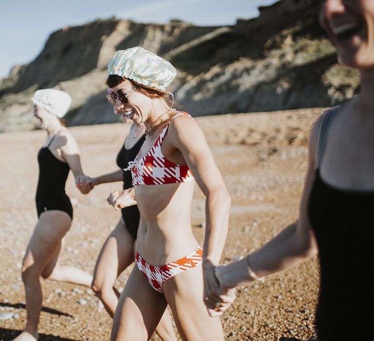 Bride in red and white check swimsuit and shower caps holds hand with friends as she runs into the sea