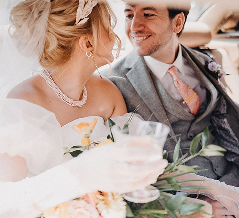 Groom wearing three piece light grey wedding suit and pink tie sitting in the back of the wedding car with the bride wearing a headpiece 