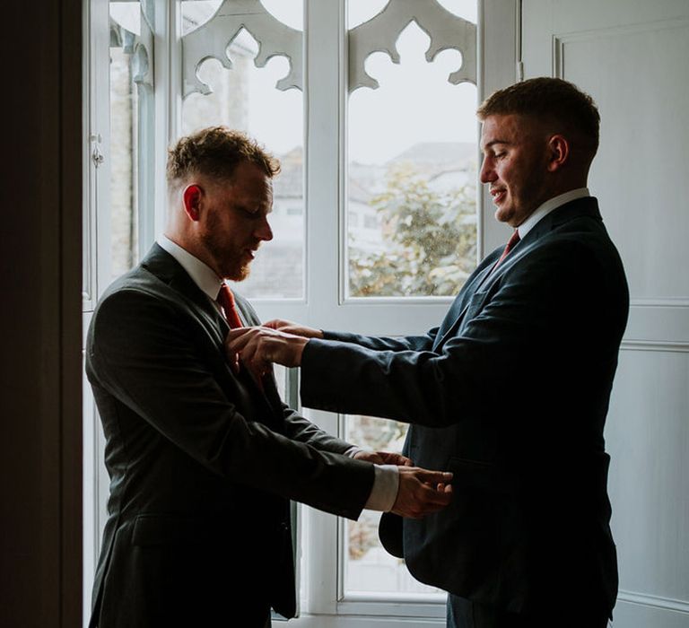 The groomsman helps the groom with his suit as the groom does up his cufflinks 