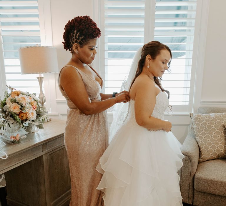 Black bridesmaid in a gold shimmer dress helping the bride put on her strapless wedding dress with full skirt 