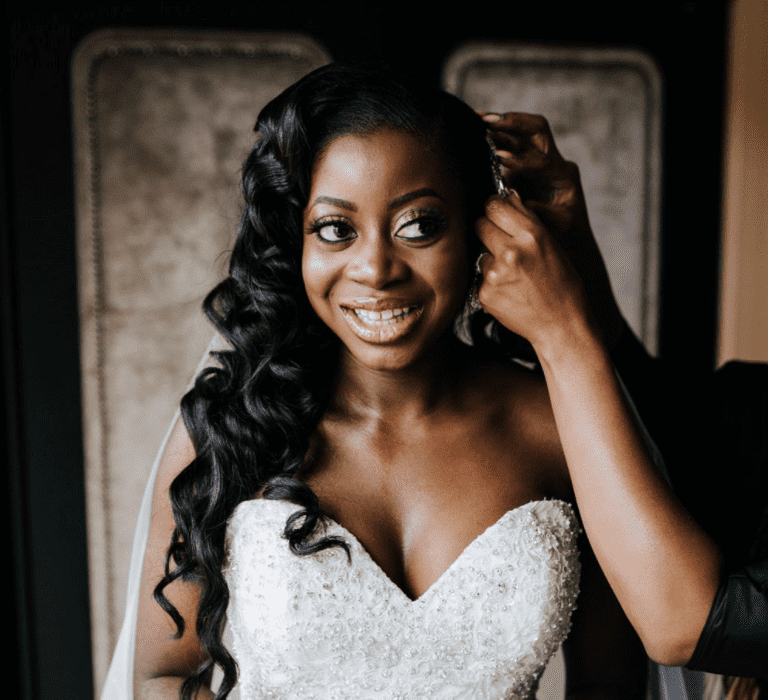 A Black bride with waist length relaxed curly hair smiles to camera as she has a clip added to her hair