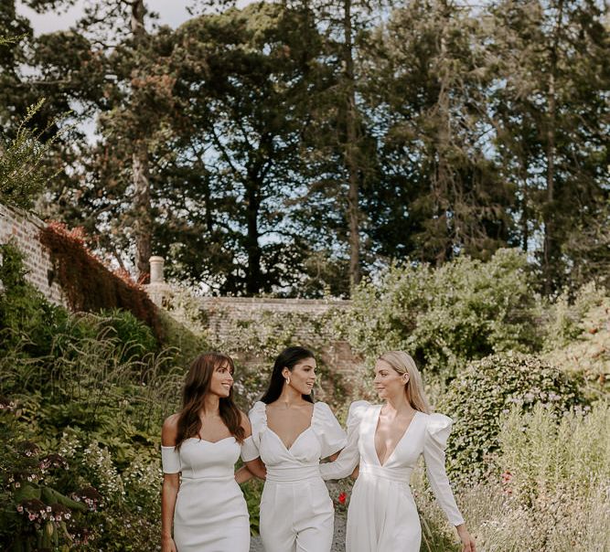 Bridesmaids in all white jumpsuits and dresses walking through the gardens at Dewsall Court