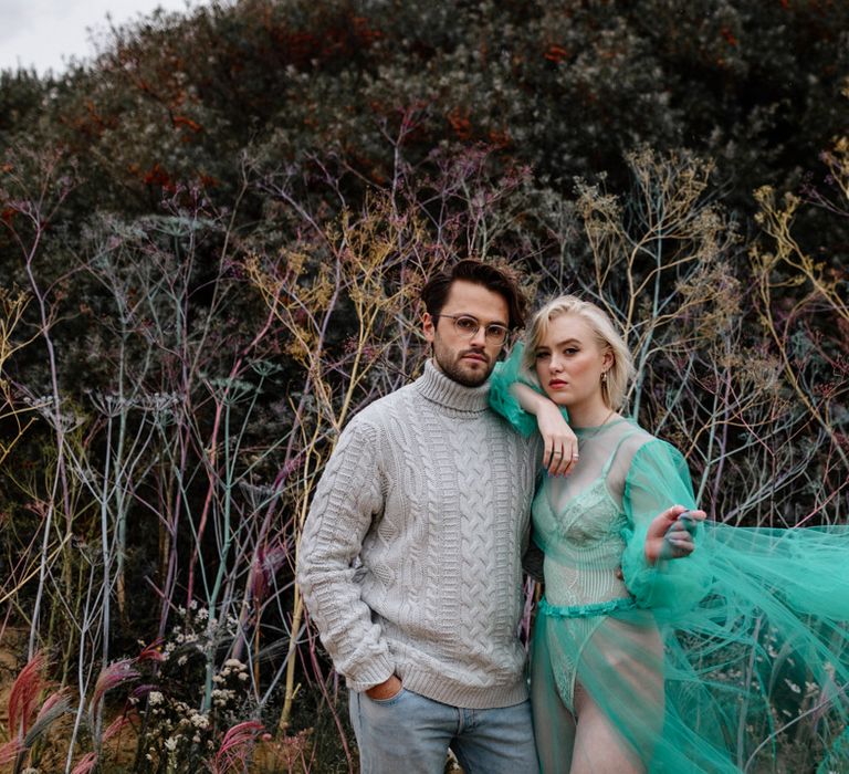 Brunette bearded groom in glasses, sweater and jeans with his bride in a sheer tulle wedding dress on the beach 