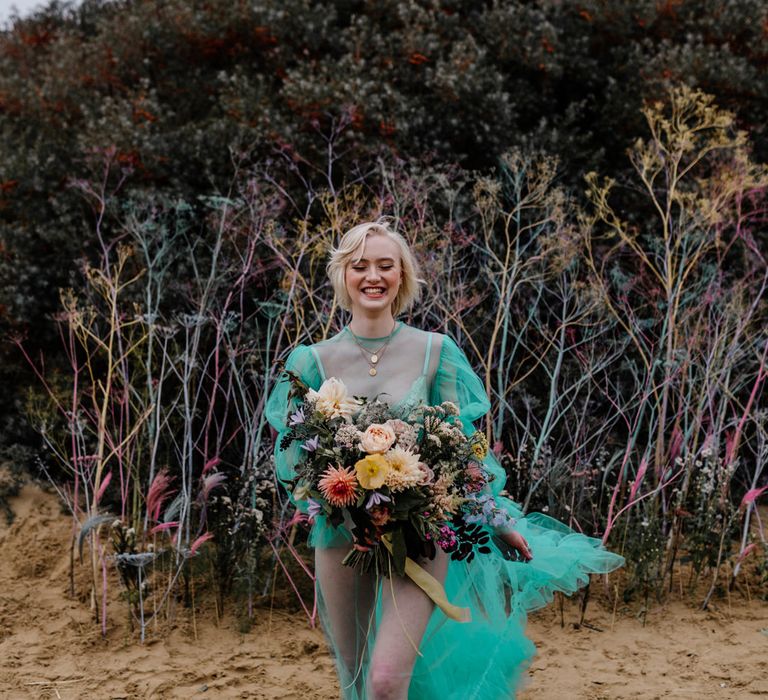 Barefoot bride walking in the sand at her beach elopement in a pastel wedding dress holding a colourful flower wedding bouquet tied with ribbon