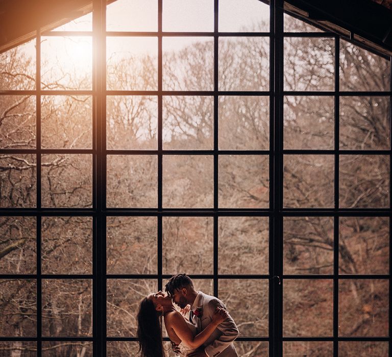 Bride & groom embrace in front of window as the sun sets behind them