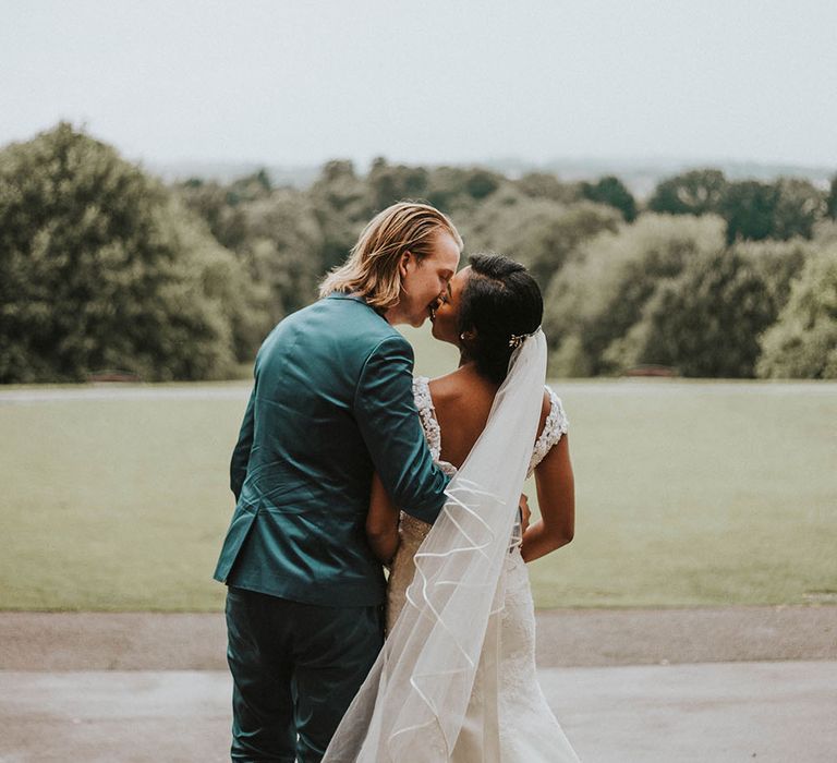 Groom in a blue suit kissing his bride in a fitted lace wedding dress and satin trimmed veil