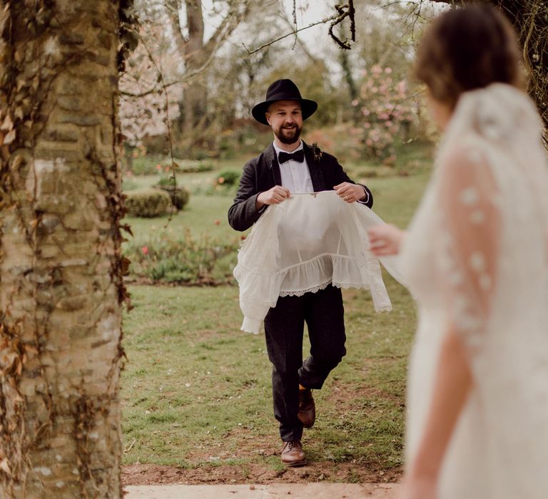 Groom in grey tweed suit, bow tie and black fedora brings bride in Charlie Brear wedding dress and applique veil her white blouse at garden party wedding in Devon