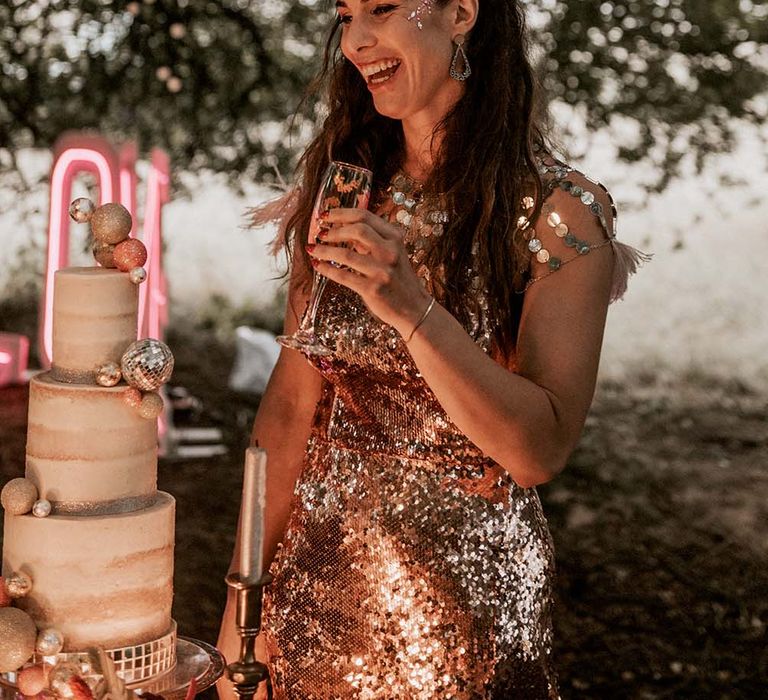 Laughing bride with long curled hair, face glitter and rose gold sequinned wedding dress holds tropical print champagne flute at festival themed wedding