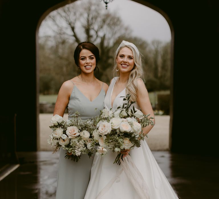 Bride stands with bridesmaid who wears sage green bridesmaids dress with V-shaped front and holds white floral bouquet