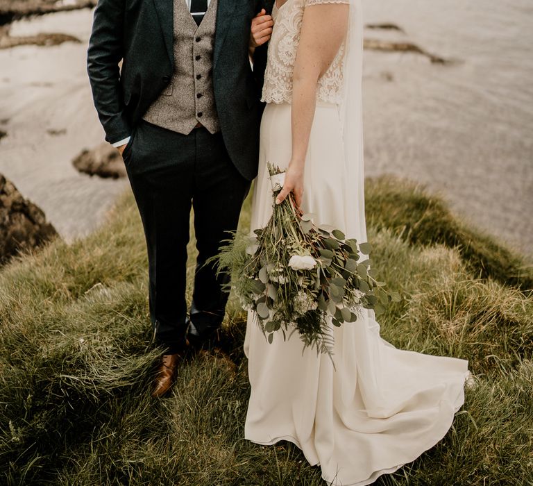 Smiling bride in lace top capped sleeve wedding dress stands with arms around groom in navy suit on clifftop after Dunluce Castle wedding