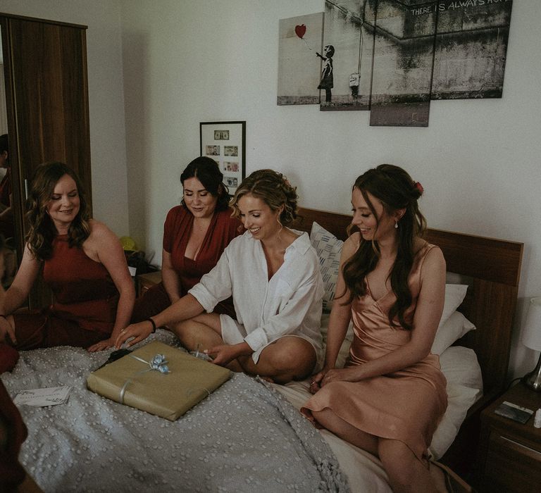 Bride in oversized white shirt sits on bed with bridesmaids in mismatched red and peach bridesmaids dresses opening present before garden wedding with burnt orange wedding theme