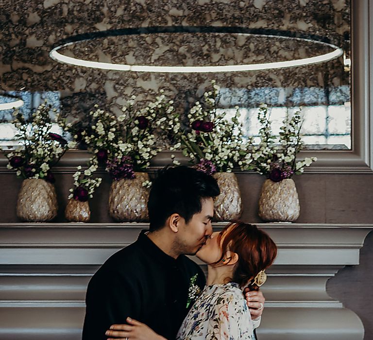 East Asian groom kissing his bride in a short floral wedding dress in front of a fireplace at Old Marylebone Town Hall 