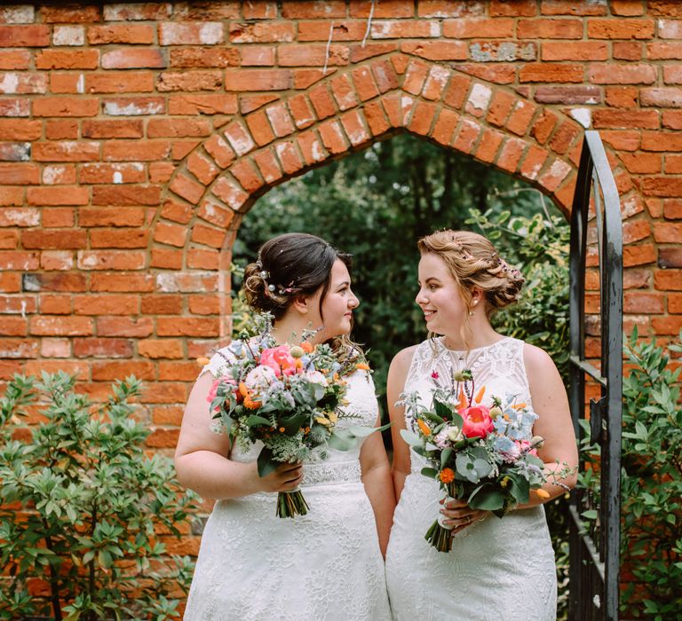 Brides look lovingly at one another as they hold bright floral bouquets before wedding ceremony