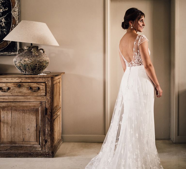 Bride with wedding updo in open back Rime Arodaky wedding dress with train looks out of the window before summer wedding in Dorset