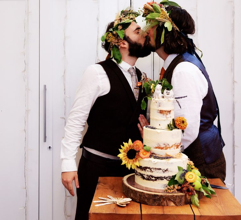 Grooms kiss in front of naked wedding cake on their wedding day