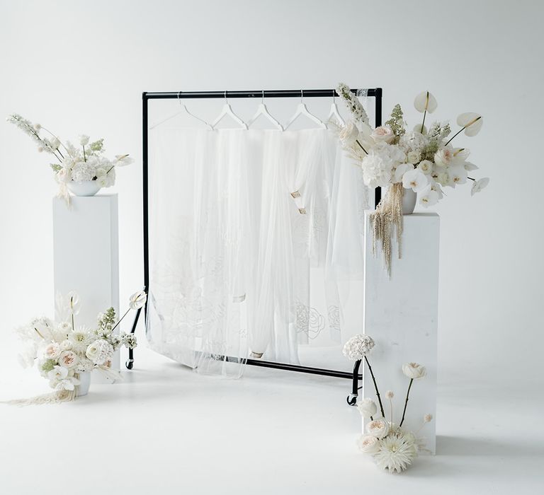 Black frame hanging rail with embroidered personalised veil ideas by Rebecca Anne Designs surrounded by all white wedding flower arrangements 