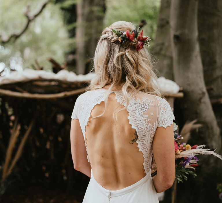 Bride in white lace cap sleeved open back wedding dress with blonde curls and flowers in her hair stands in woodland at late summer wedding in Norfolk