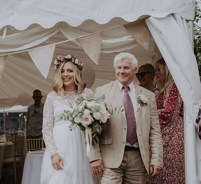 Father of the bride in a beige suit and pink tie escorting his daughter in a lace and tulle wedding dress with long sleeves to the back garden wedding ceremony 