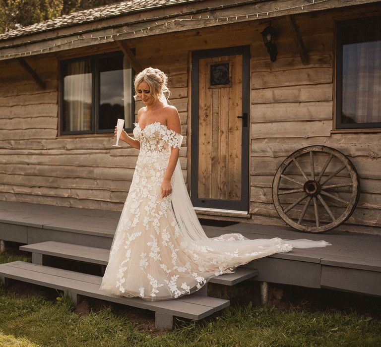 Bride in off the should Enzoani wedding dress with train and veil holds glass of sparkling wine as she walks down wooden steps before tipi wedding at Inkersall Grange Farm