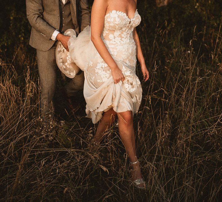 Bride in white off the shoulder Enzoani wedding dress and veil walks through grass as groom in grey three piece suit and white tie holds the train of her dress at Inkersall Grange Farm wedding