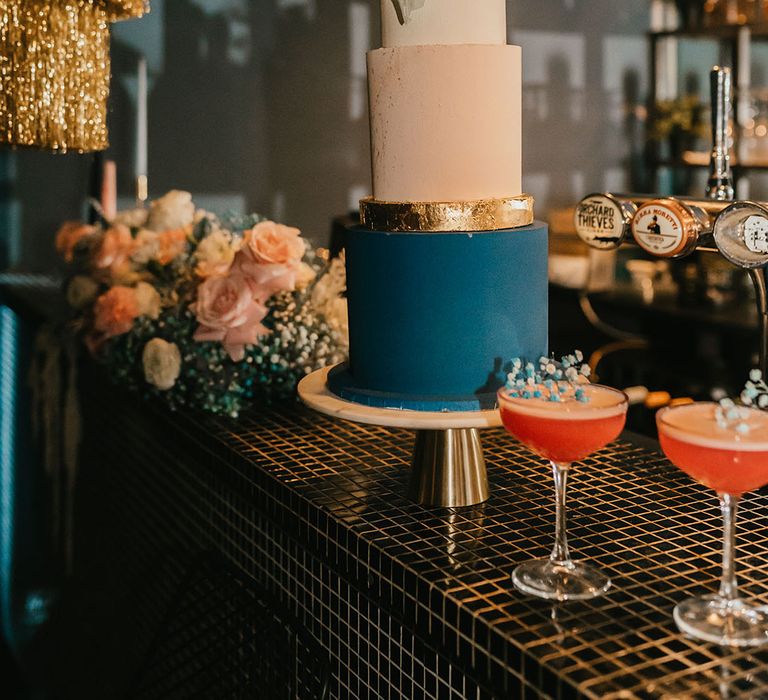 White, light pink, gold and teal wedding cake with sugar shard cake topper resting not the bar with colourful cocktails 