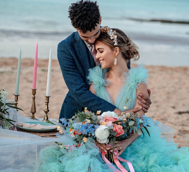 Groom in a blue wool suit embracing his bride in a mint green tulle wedding dress as she holds a pastel wedding bouquet tied with ribbon 