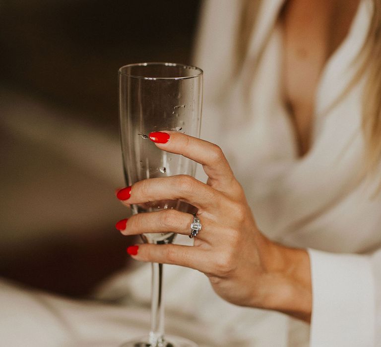 Bride holds champagne flute and wears rectangle diamond engagement ring with bright pink nail varnish 