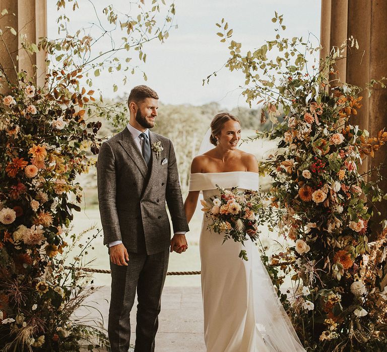 Bride & groom stand under floral archway outdoors in front of large pillars as they hold hands