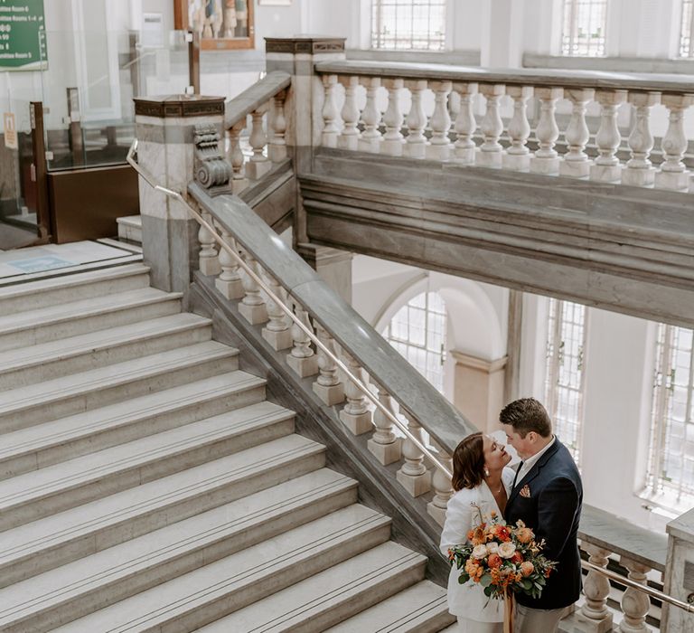 Bride in a trouser suit holding an orange flower bouquet and groom in a blue blazer at Islington Town Hall wedding 