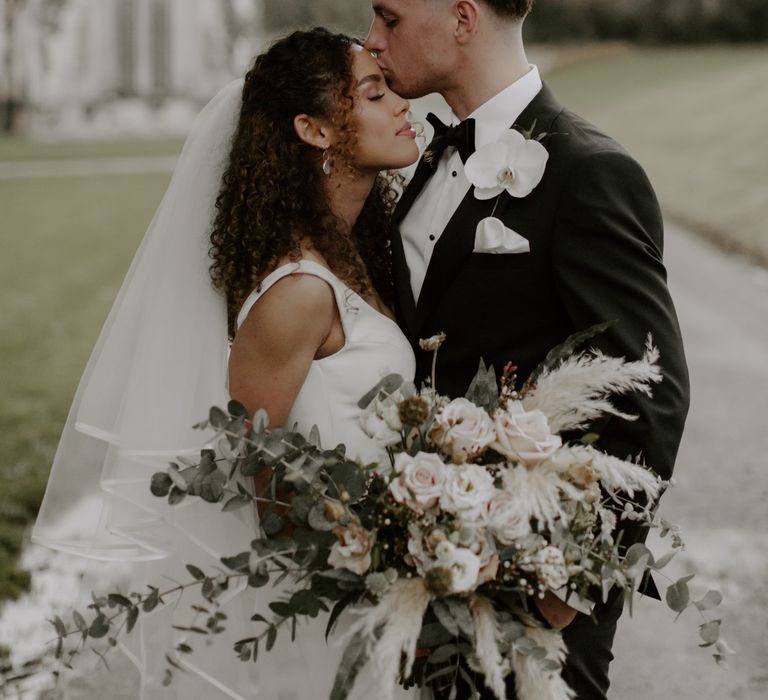 Groom kisses the top of his brides forehead as they stand outdoors on their wedding day