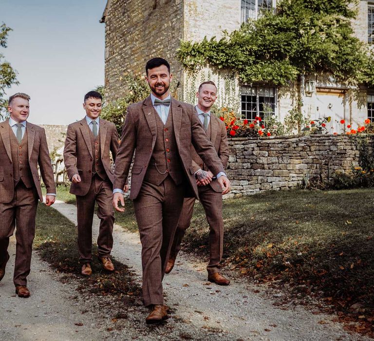 Groom and groomsmen all in brown three piece tweed suits with orange buttons walk down driveway for Costwolds wedding