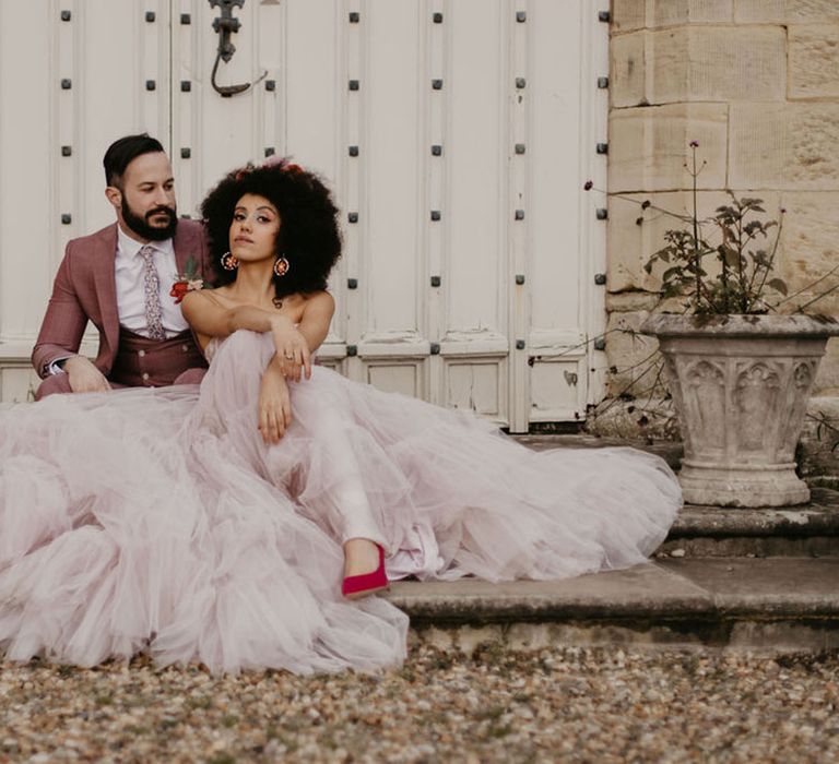 Black bride in a pink tule full skirt and red velvet stiletto wedding shoes sitting on the steps at Chiddingstone Castle with her groom in a pink suit 