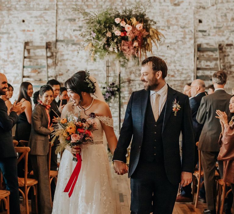 Bride in lace off the shoulder wedding dress and pearl necklace holding orange, pink and green bridal bouquet tied with red ribbon walks down the aisle with groom in dark blue three piece suit