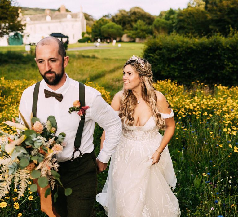 Groom leads the bride through field of wildflowers with autumnal wedding bouquet