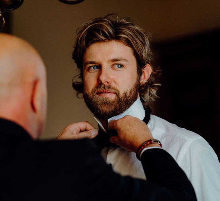 Bearded groom with long wavy hair putting his bow tie on 