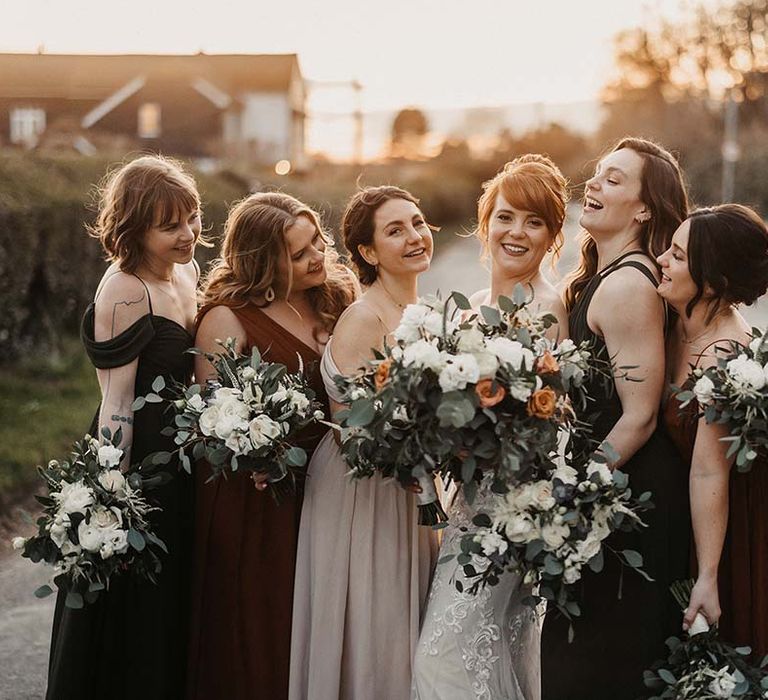 Bride laughs with her bridesmaids in earth toned dresses with various necklines outside barn wedding venue 