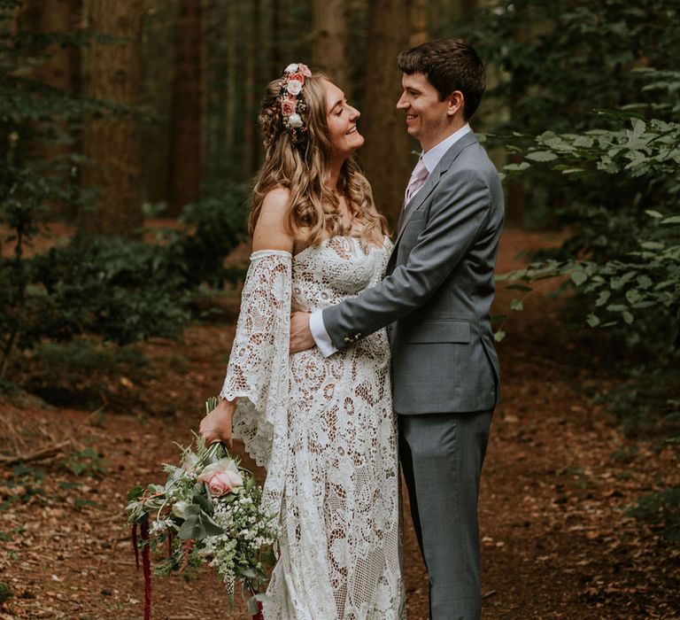 Groom in grey suit and pink tie hugs bride in lace medieval wedding dress and flower crown holding pink rose andwhite gypsophila bouquet