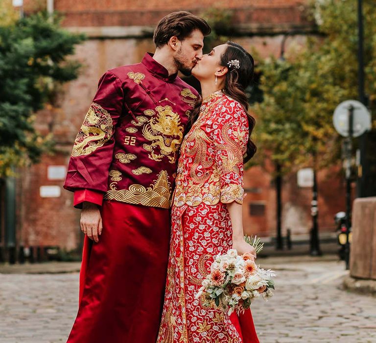 Bride with curled sideswept hair and accessory in traditional Chinese wedding attire kisses groom in matching attire for multicultural wedding