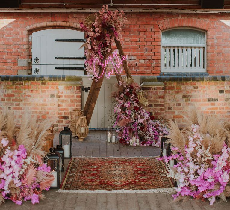 Rustic wedding styling with wooden benches, wool rugs, lanterns and wooden frame altar decorated with a pink neon sign, dried grasses and pink flowers