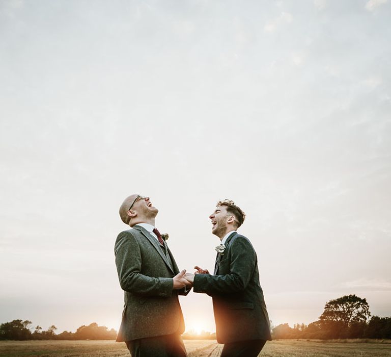 Grooms wearing matching red ties laugh together in a field as the sun starts to set |