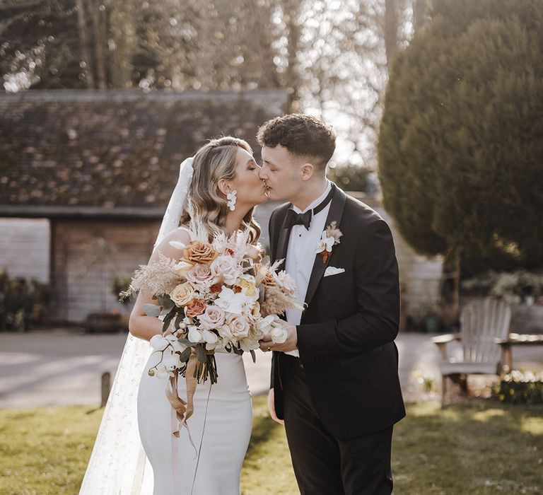 Bride holding neutral wedding bouquet with roses, orchids and dried flowers and flower earrings kisses groom in black tie with white flower buttonhole